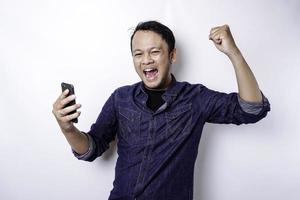 ein junger asiatischer mann mit einem glücklichen erfolgreichen ausdruck, der blaues hemd trägt und sein telefon hält, lokalisiert durch weißen hintergrund foto