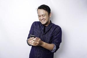 Aufgeregter asiatischer Mann mit blauem Hemd lächelt, während er sein Telefon hält, isoliert durch weißen Hintergrund foto