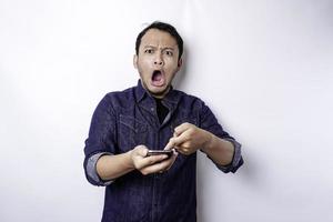 schockierter asiatischer mann, der blaues hemd trägt und sein telefon hält, isoliert durch weißen hintergrund foto