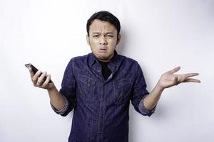 ein nachdenklicher junger asiatischer mann trägt ein blaues hemd, das sein telefon hält, und sieht verwirrt aus, isoliert durch weißen hintergrund foto