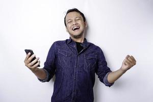 ein junger asiatischer mann mit einem glücklichen erfolgreichen ausdruck, der blaues hemd trägt und sein telefon hält, lokalisiert durch weißen hintergrund foto