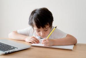 Grundschulkind, Junge, der das Schreiben lernt, Hausaufgaben zu Hause sitzend, Porträt-Vorschulkind, das online studiert, Notiz mit Bleistift macht, indem er Computer für seine Hausaufgaben, Bildung und Technologiekonzept verwendet foto