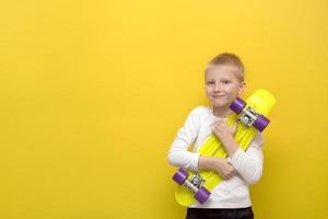 Blonder Junge mit Überraschung und Freude kuschelt ein Skateboard auf gelbem Hintergrund mit Kopierraum, ein Geschenk foto