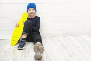 Junge mit Hut sitzt auf dem Boden in der Nähe der Wand mit einem Skateboard in den Händen, aktiver Lebensstil foto