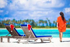 weihnachtskonzept, strandliegen mit roten weihnachtsmützen und schönes mädchen während des tropischen urlaubs foto