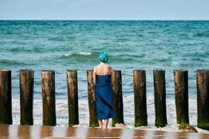 junge blauhaarige frau in langem dunkelblauem kleid, die am sandstrand steht und den ozeanhorizont betrachtet foto