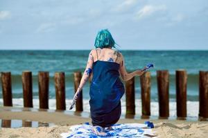 Künstlerische blauhaarige Performance-Künstlerin mit blauen Gouache-Farben, die am Strand tanzen foto