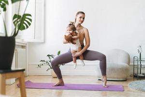 junge frau passte mutter mit babymädchen in ihren armen, die zu hause fitness auf der matte machen foto