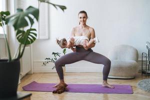 junge frau passte mutter mit babymädchen in ihren armen, die zu hause fitness auf der matte machen foto