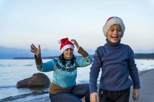glückliche mutter und kleiner sohn feiern weihnachten am strand in weihnachtsmützen, der junge lacht fröhlich, unschärfe foto