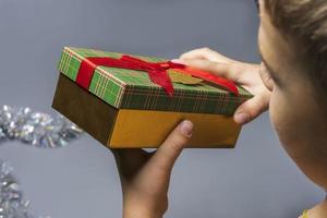 Ein Junge mit einem Weihnachtsgeschenk in den Händen auf grauem Hintergrund, eine Schachtel mit einem Neujahrsgeschenk in den Händen eines Kindes foto