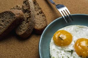 Spiegeleier auf einem türkisfarbenen Teller, bestreut mit Dill, Gabel und Brotstücken, Draufsicht, Frühstück mit Spiegeleiern foto