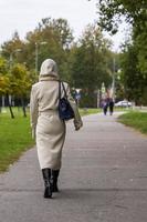 Ein Mädchen in einem modischen beigefarbenen Herbstmantel, Stiefel mit einer modischen Tasche geht auf einem Weg in einem Stadtpark spazieren foto