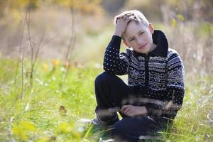 ein hübscher ruhiger junge in einem strickpullover sitzt auf dem gras auf dem feld. foto