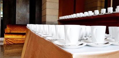 viele tassen kaffee oder tee und edelstahllöffel setzen auf einen weißen tisch, um kunden und personal im hotel oder ballsaal zu bedienen. gruppe von objekten, essen, trinken, service und vorbereitungskonzept foto