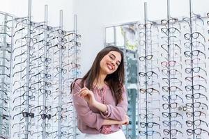 gesundheitswesen, sehvermögen und visionskonzept - glückliche frau, die sich im optikgeschäft für eine brille entscheidet. junges schönes mädchen optikgeschäft, das neue brille anprobiert foto