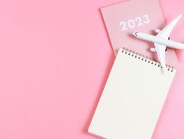Flaches Notizbuch aus leerem Papier auf rosafarbenem Tagebuch 2023 und Flugzeugmodell auf rosafarbenem Hintergrund. Reiseplan-Konzept. foto