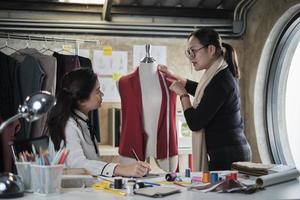 asiatische modedesignerin mittleren alters unterrichtet junge jugendliche schneiderin über form und größe mit maßband und nähstoff für kleiderdesign-kollektion, professionelles boutique-kleinunternehmen. foto