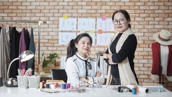 Modeteam, asiatische Designerin und jugendliche Assistentin im Studio, verschränkte Arme und Lächeln, glückliches Arbeiten mit bunten Fäden und Nähen für Kleiderdesign, professioneller Boutique-Schneider, kleiner Unternehmer. foto