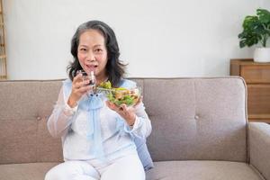 asiatische Seniorin, die Gemüsesalat und gesundes Essen isst und glücklich auf dem Sofa im Haus für einen gesunden Körper isst. gesundes lebensmittelkonzept foto