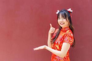 süßes asiatisches mädchen mit langen haaren, das ein rotes cheongsam-kleid im chinesischen neujahrsthema trägt, während sie ihre hand zeigt, um etwas auf rotem hintergrund zu präsentieren. foto