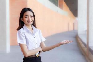 asiatische schöne junge studentin lächelt und schaut in die kamera, die steht, um etwas selbstbewusst im universitätshintergrund zu präsentieren foto