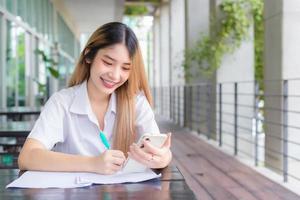 junge asiatische studentin in uniform, die smartphone benutzt und etwas über die arbeit schreibt. es gibt viele dokumente auf dem tisch, ihr gesicht mit einem lächeln in einem arbeiten, um informationen zu suchen. foto