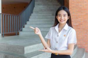 süße asiatische junge studentin lächelt und schaut in die kamera, die steht, um etwas selbstbewusst im universitätshintergrund zu präsentieren. foto