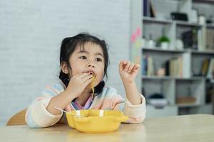 Asiatisches Mädchen glücklich mit Bestecklöffel und Gabel, das köstliche Nudeln und Fleischbällchen in der Küche am Esstisch isst. Fröhliches asiatisches Mädchen übt, alleine am Esstisch zu essen. Babynahrungskonzept foto