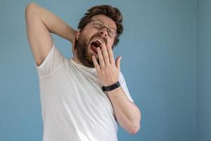 bärtiger mann mit brille und weißem t-shirt gähnt mit weit geöffnetem mund und bedeckt ihn mit der hand, er ist schläfrig oder gelangweilt. Blauer Hintergrund. foto