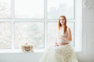 Rothaariges schwangeres junges Mädchen in einem weißen Kleid in der Nähe des Fensters foto