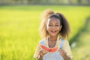 schwarzhäutiges süßes kleines Mädchen, das im Freien Wassermelone isst grüne Reisfeldkulisse afrikanisches Kind, das Wassermelone isst foto