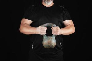 Erwachsener starker Athlet in schwarzer Kleidung, der eine eiserne Kettlebell auf seinen ausgestreckten Armen hält foto