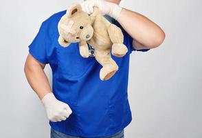 Arzt in blauer Uniform, der als Zeichen der Verzweiflung einen Teddybären mit Pflaster am Kopf hält foto