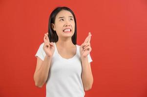 porträt einer jungen besorgten asiatischen frau im weißen t-shirt, die die daumen für viel glück drückt und den großen wunsch zum ausdruck bringt, isoliert auf rotem hintergrund zu gewinnen foto