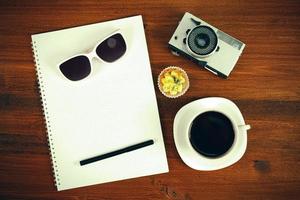 retro film fotokamera, sonnenbrille, cupcake, tasse kaffee und notizbuch mit bleistift auf braunem tisch. Ansicht von oben. foto