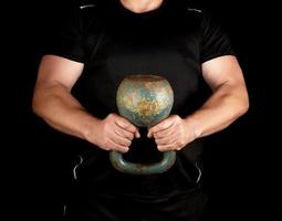 Erwachsener starker Athlet in schwarzer Kleidung, der eine eiserne Kettlebell auf seinen ausgestreckten Armen hält foto
