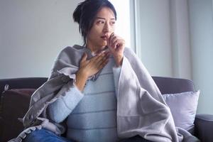 asiatische Frau ist krank mit verstopfter Nase, fließendem Wasser in den Händen, Wischpapier, hohem Fieber, das auf dem Sofa im Haus sitzt foto
