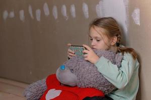 ein trauriges neunjähriges Mädchen, sie sitzt in einem Zimmer auf dem Dachboden ohne Reparaturen auf dem Boden, sie umarmte einen alten großen Teddybären, sie hat ein Telefon in der Hand, sie kommuniziert in sozialen Netzwerken foto