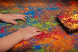 Kinderhände liegen auf einem Tisch, auf dem Stechpalmenfarben verstreut sind, und eine Holzplatte in Form eines Herzens, in der leuchtende Farben Platz kopieren. indisches fest der farben holi. foto