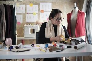 asiatische modedesignerin mittleren alters arbeitet im studio, schneidet und wählt stoff- und fadenfarben mit zeichnungsskizzen für kleiderdesign-kollektionen. professioneller Boutique-Schneider-Unternehmer. foto