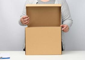 Frau in grauer Kleidung steht und hält offene braune Kiste auf weißem Hintergrund, bewegt, sendet und liefert Waren foto