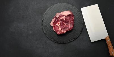 Rundes rohes Stück Rindfleisch auf einem schwarzen Brett. Ribeye zum Grillen foto