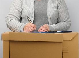 frau in grauer kleidung steht neben einer braunen kiste und unterschreibt dokumente mit einem stift. das Konzept des Bewegens, Sendens und Empfangens von Waren foto