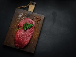 rohes rinderfilet liegt auf einem schneidebrett und gewürzen zum kochen auf einem schwarzen tisch, draufsicht foto