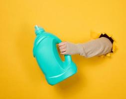 Blaue Plastikflasche mit Flüssigwaschmittel in weiblicher Hand auf gelbem Hintergrund. ein Körperteil ragt aus einem ausgerissenen Loch im Hintergrund foto