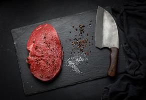 rohes rinderfilet liegt auf einem schneidebrett und gewürzen zum kochen auf einem schwarzen tisch, draufsicht foto