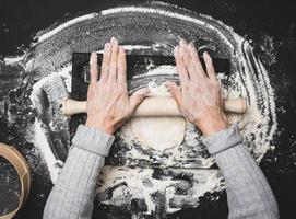 Zwei weibliche Hände rollen weißen Weizenmehlteig mit einem hölzernen Nudelholz auf einem schwarzen Tisch aus, Draufsicht foto