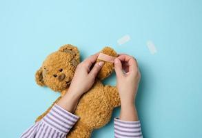 Die weibliche Hand hält einen braunen Teddybären und klebt ein medizinisches Heftpflaster auf blauem Hintergrund, Straßenbahnbehandlung foto