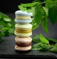 Stapel von mehrfarbigen Macarons auf schwarzem Hintergrund, hinter grünen Zweigen der Minze, foto